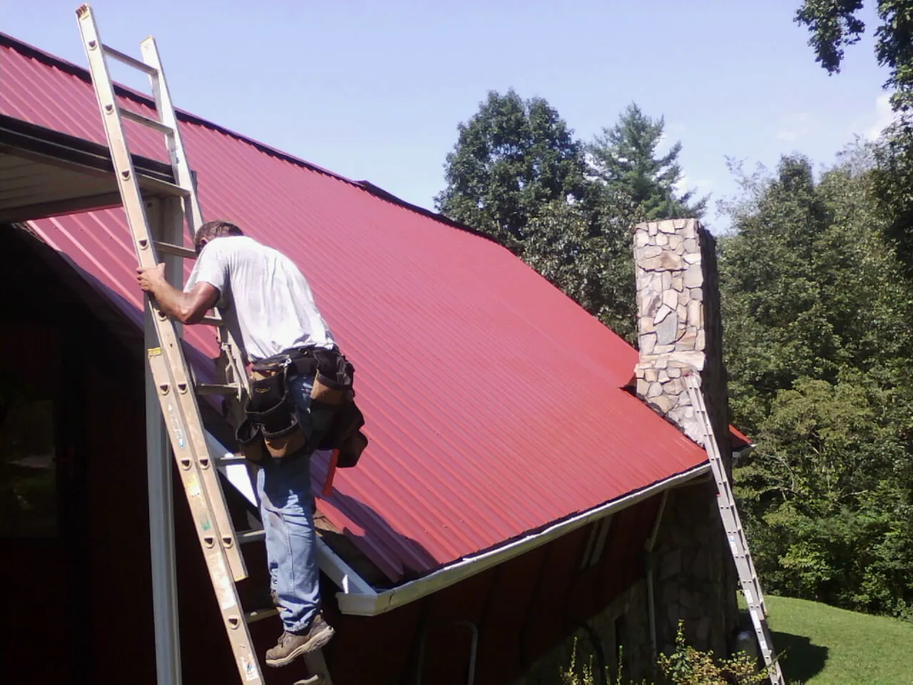 Roofer on a ladder beside a red metal roof and stone chimney — Soffit & Fascia Repair by Roofing Brothers in Dunreith